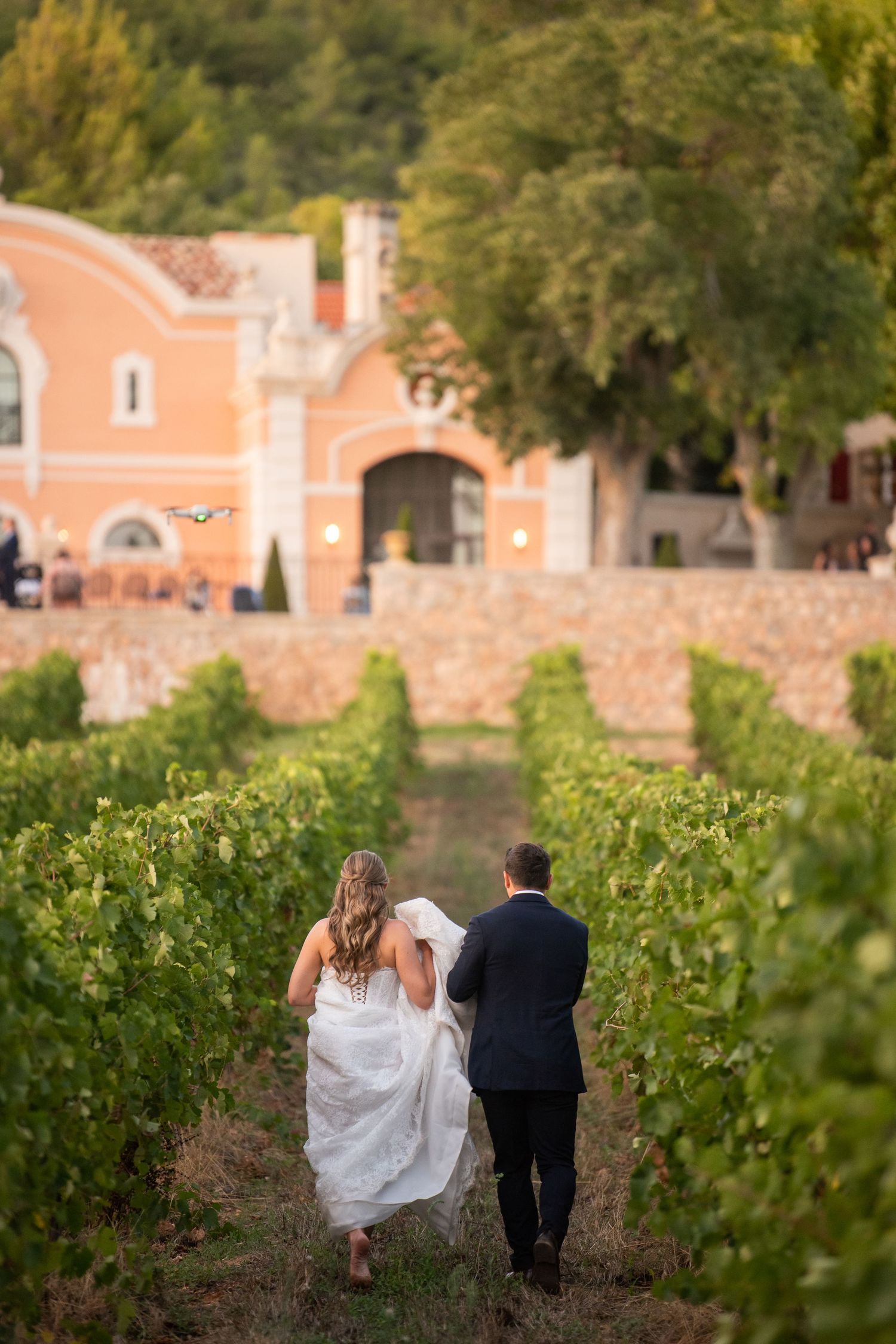 A couple walks through lush vineyard rows with rustic terracotta buildings in the background on a sunny day.