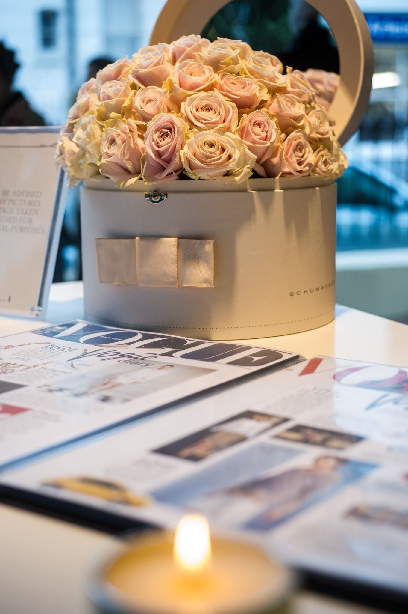 Elegant box filled with pale pink roses next to magazines and a lit candle.
