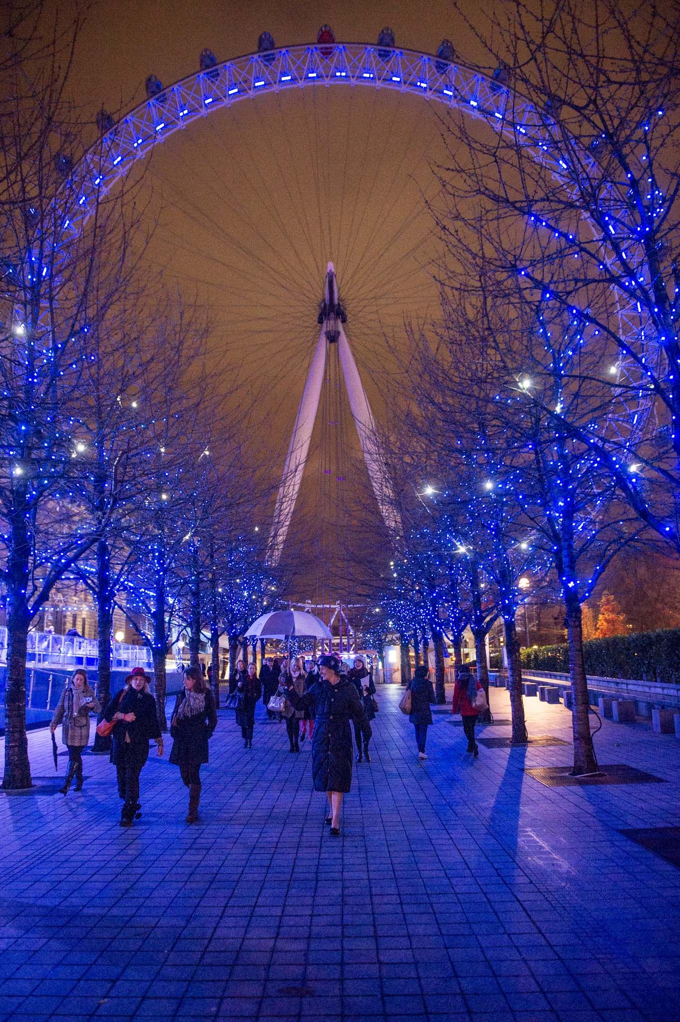 London Eye illuminated in blue at night with festive winter trees along South Bank walkway.