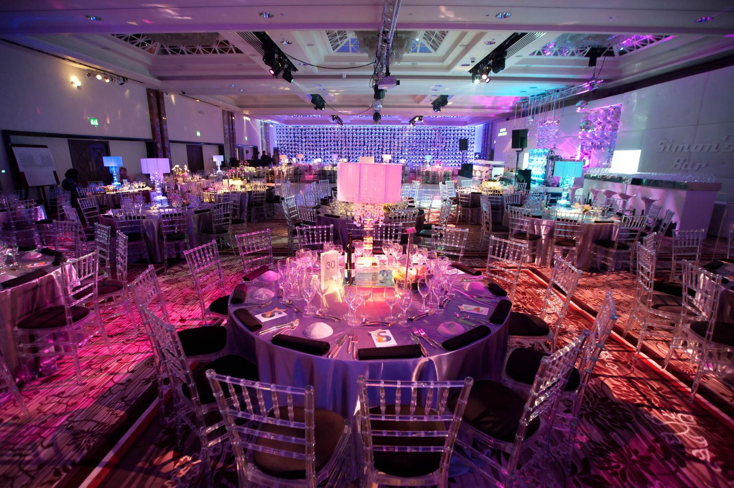 Elegant ballroom venue with purple and pink uplighting features round tables and chairs arranged in a curved formation.