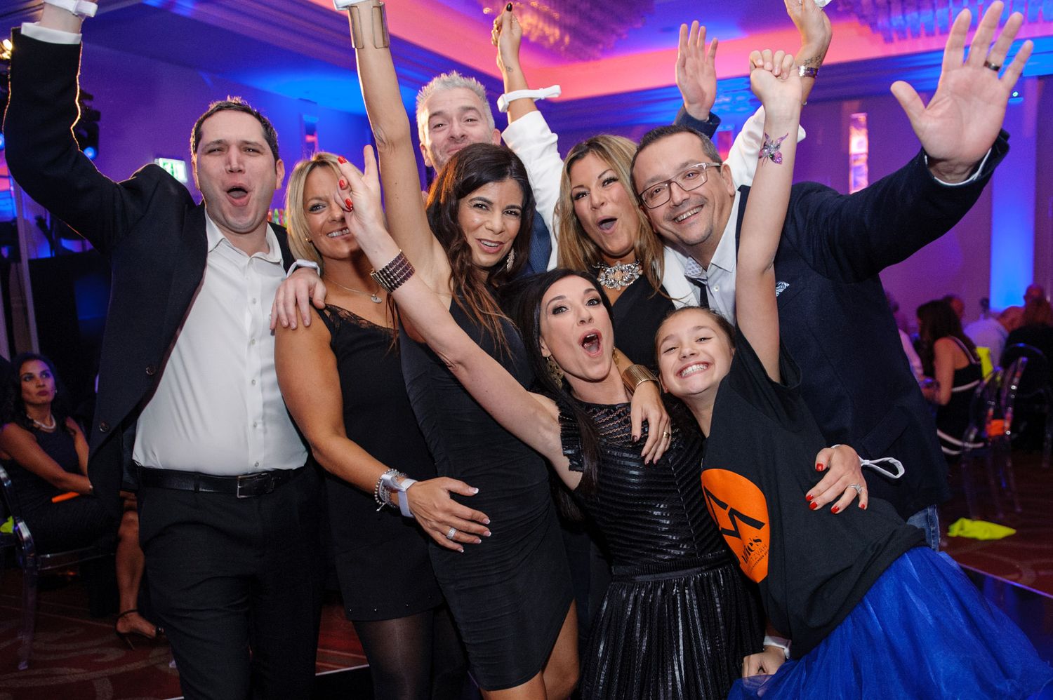 Excited wedding guests dancing and celebrating with raised arms on a colorfully lit dance floor at a reception party.
