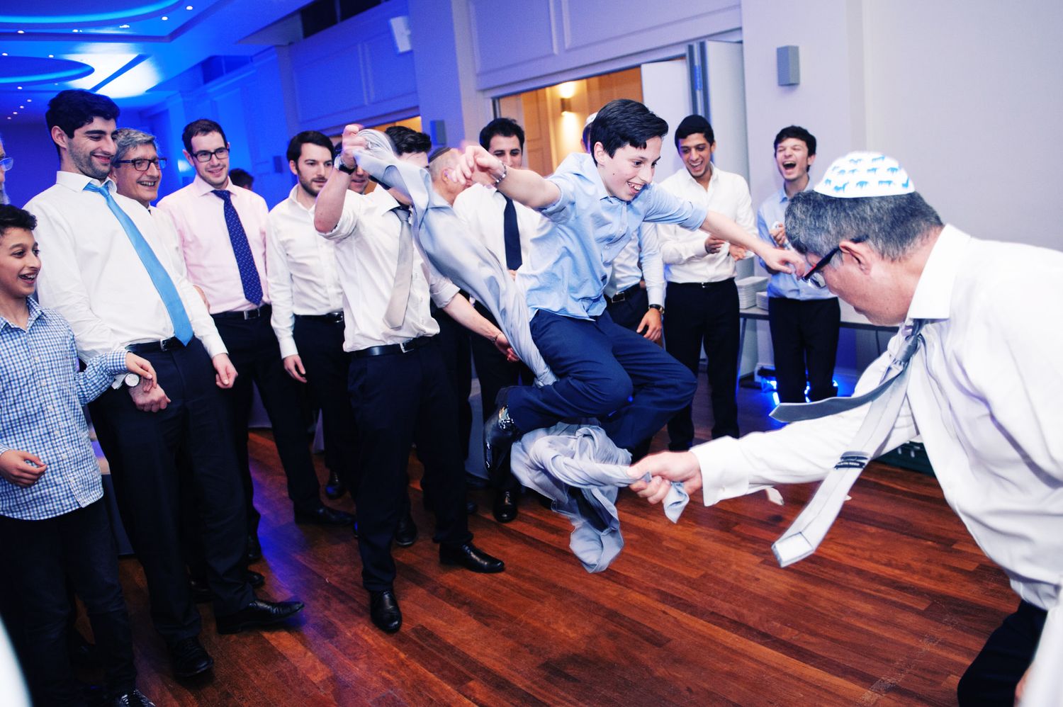 People in formal attire dancing and jumping energetically on a wooden dance floor at an indoor celebration.