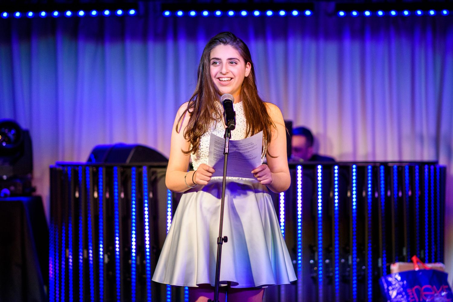 A performer in a silver dress stands on stage with blue lighting and curtains in the background.
