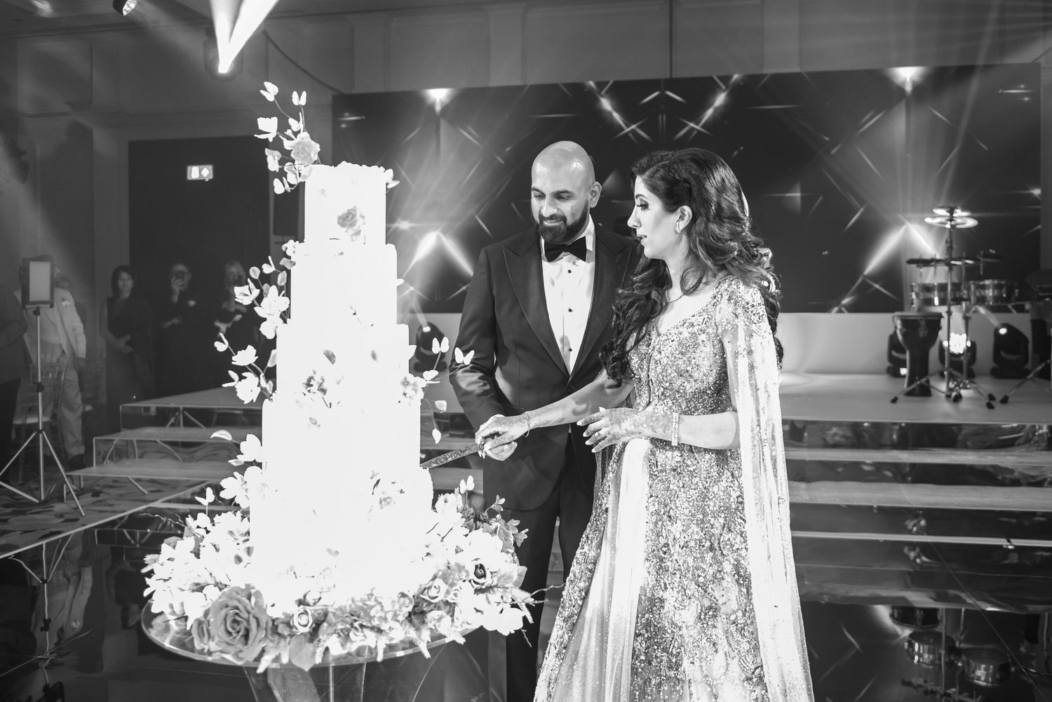 A couple in elegant wedding attire cuts into a tall, ornate white wedding cake at their reception celebration.