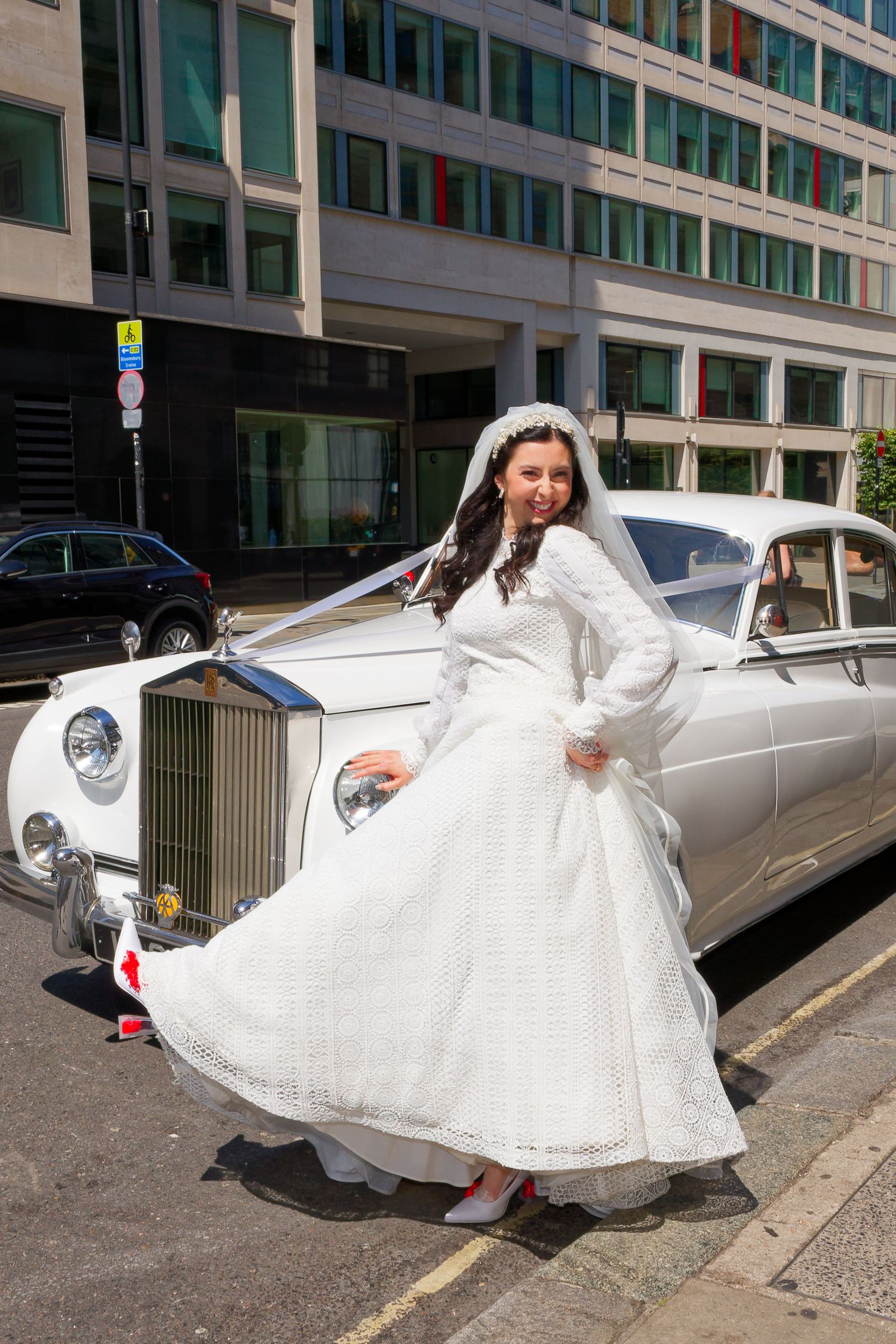 A bride in a vintage white lace dress poses elegantly next to a classic car on a city street.