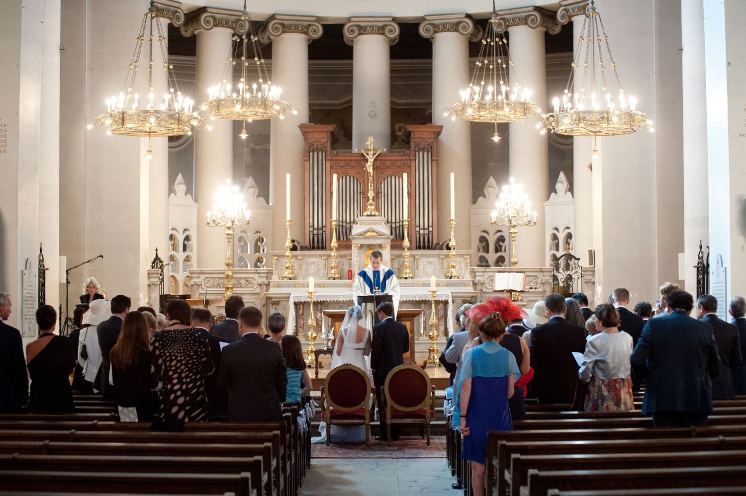 Congregation standing during a church ceremony with ornate altar, chandeliers, and clergy in blue vestments.