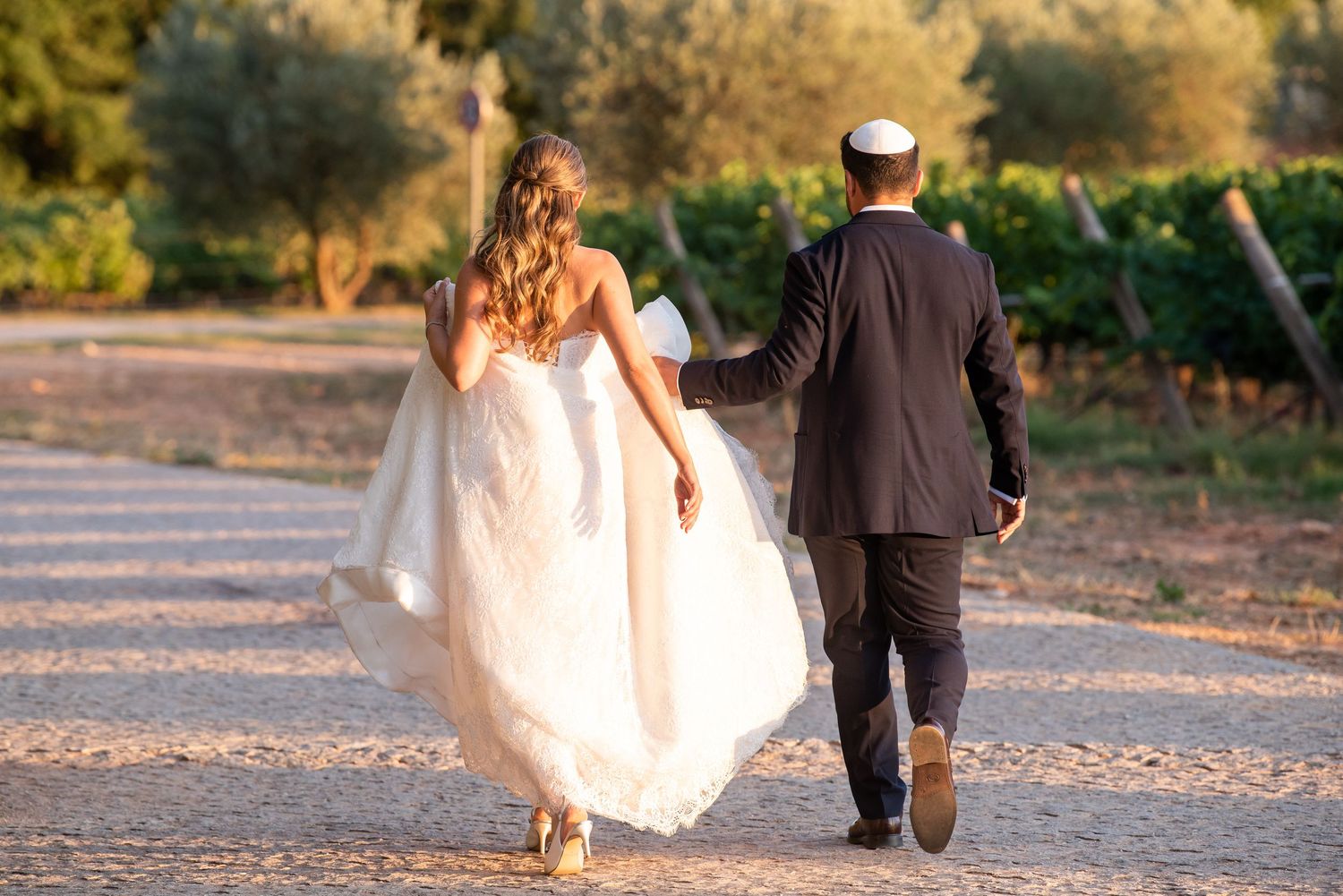 Bride and groom walking hand in hand through a vineyard at sunset, bride in white gown, groom in dark suit.