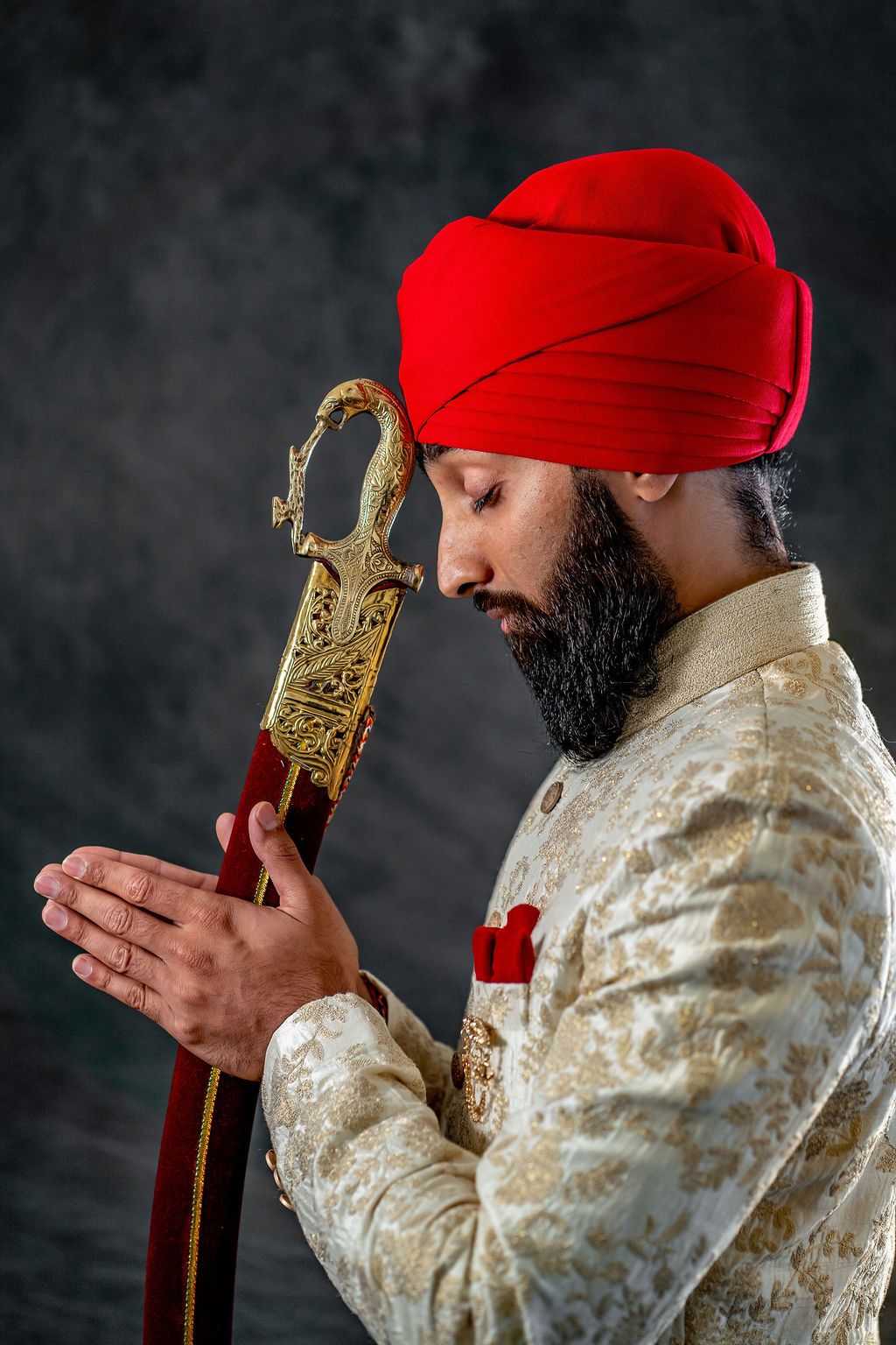 Sikh groom in red turban and gold sherwani holds ornate ceremonial sword in prayer pose against dark background.