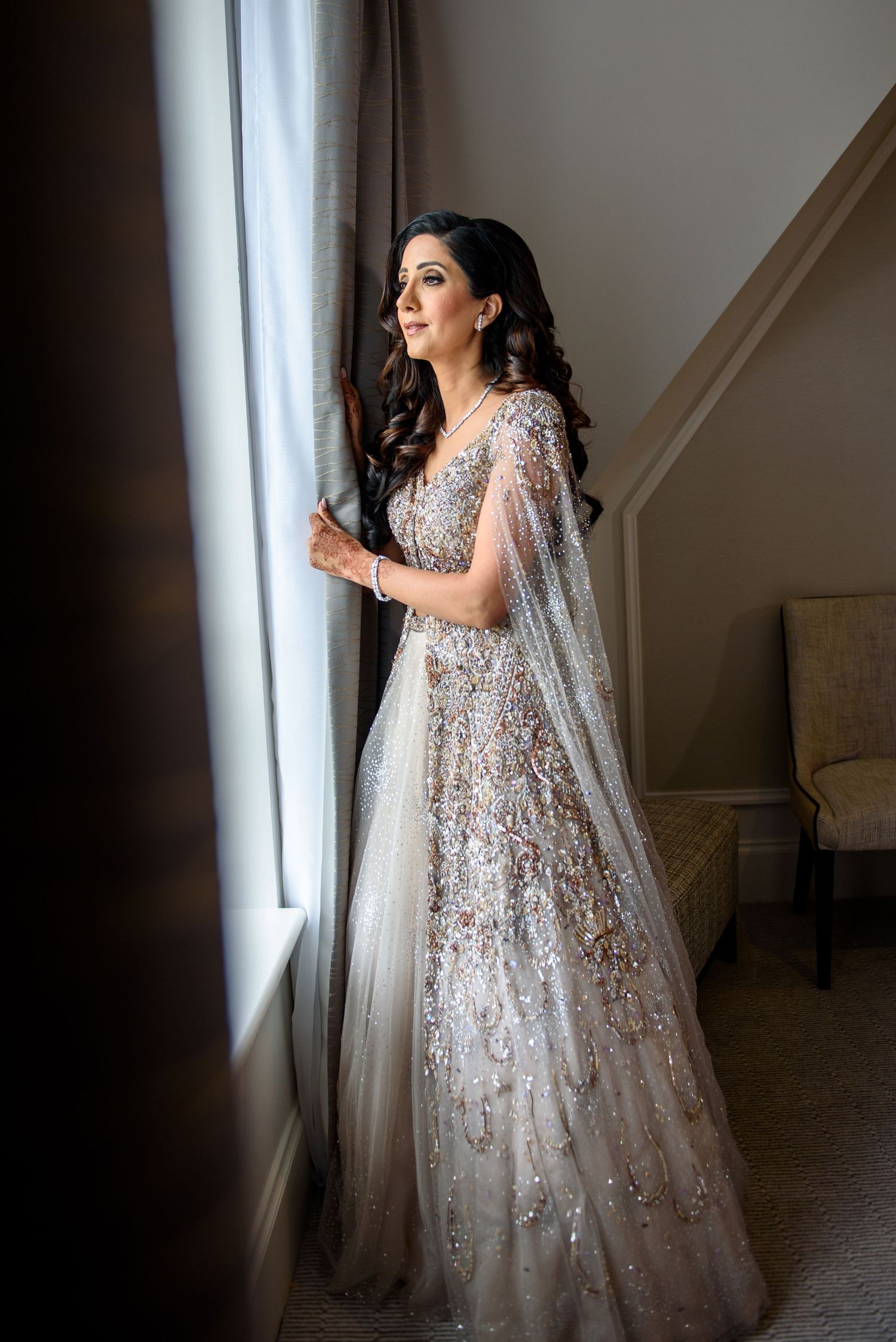 A bride in a sparkling white and silver embellished gown stands elegantly by a window in soft natural light.