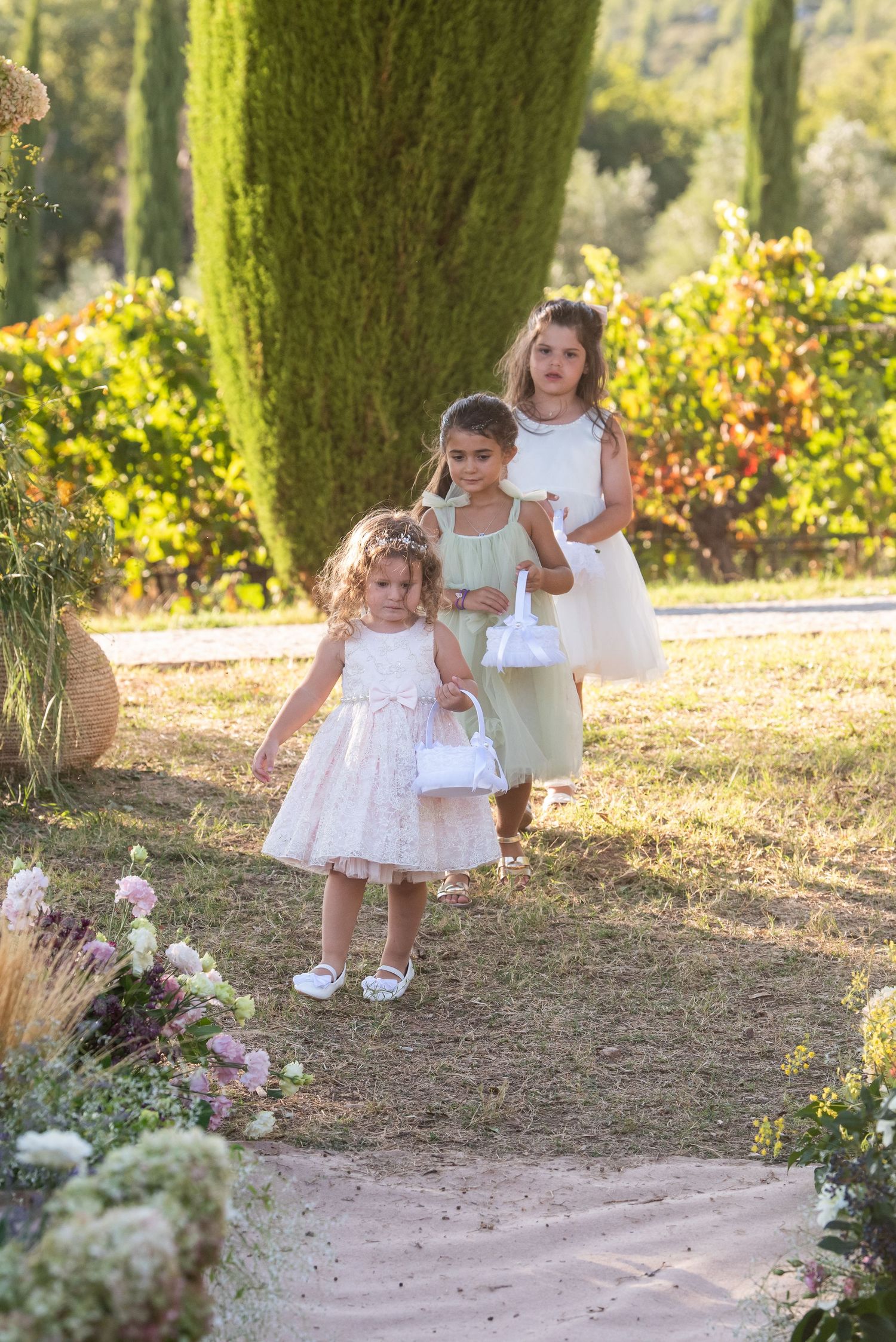 Three children in white dresses walk along a garden path surrounded by lush greenery and flowers on a sunny day.