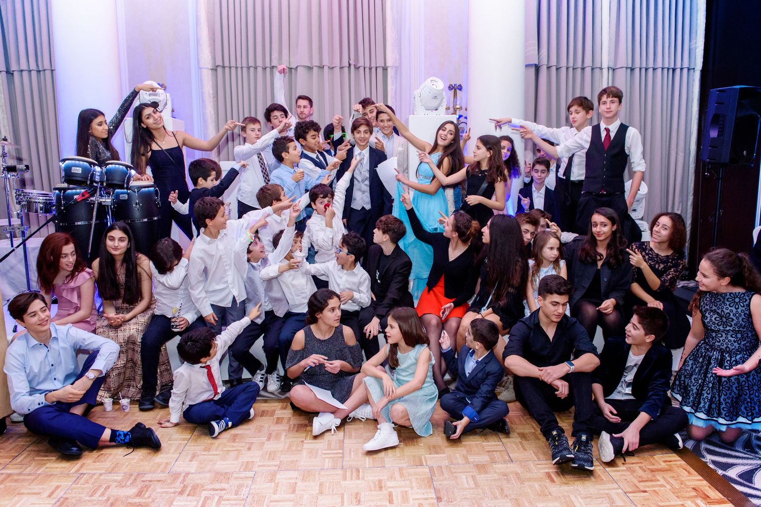 A large group of people sitting together on the floor against a wall during an indoor event or gathering.