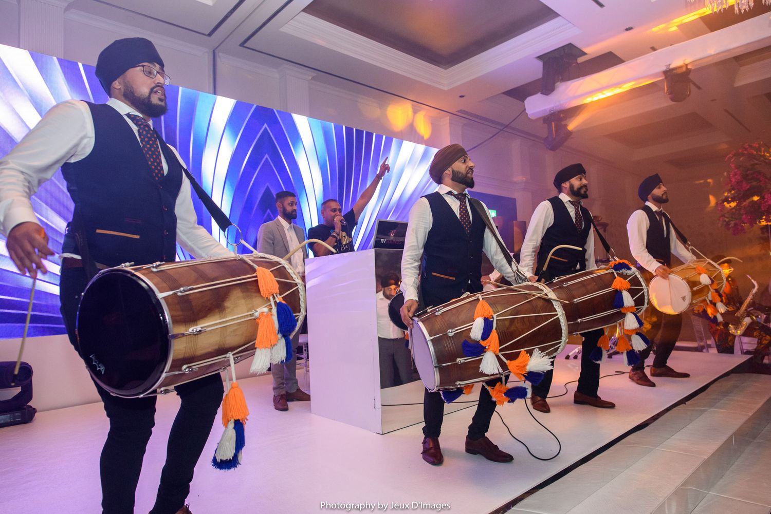 Sikh dhol drummers in traditional attire performing at a festive wedding celebration in an elegant banquet hall.