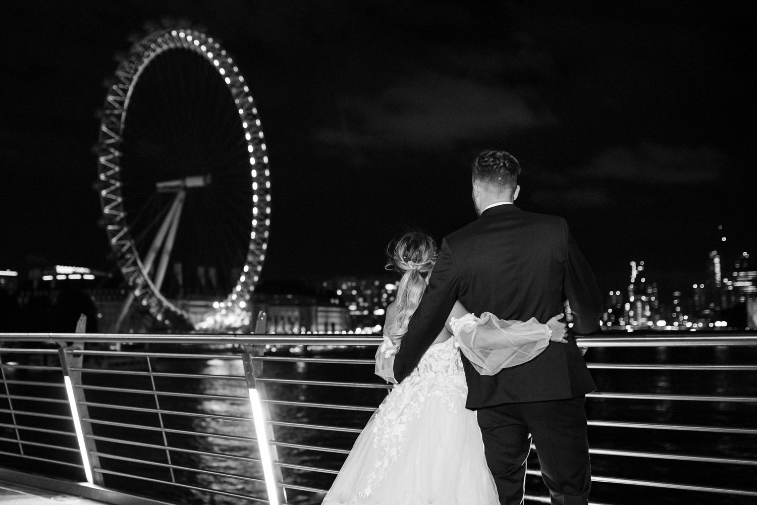 Black and white photo of a bride and groom embracing with the London Eye illuminated at night in the background.