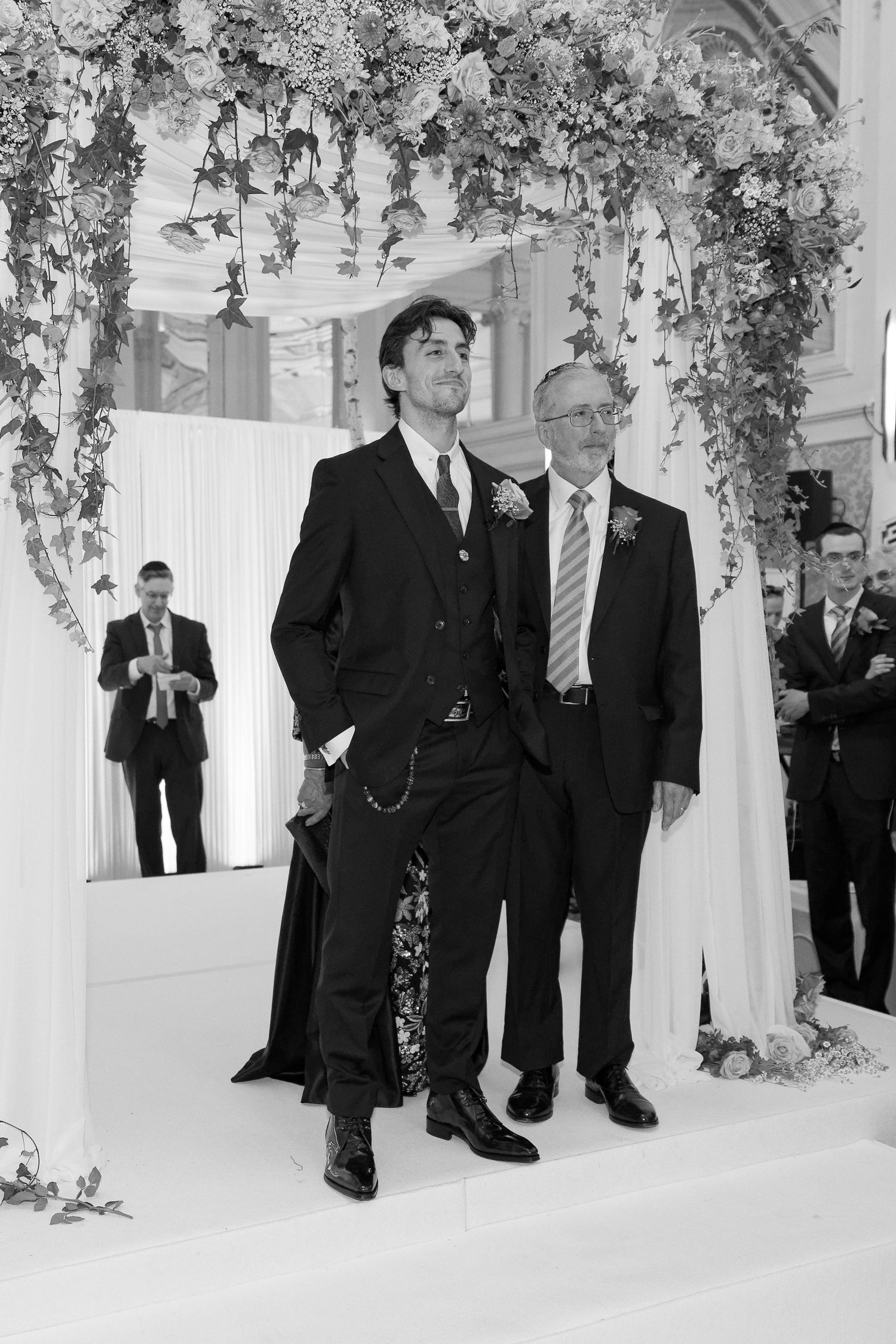 A group of men in dark suits stand beneath floral decorations in an elegant white room during a formal event.
