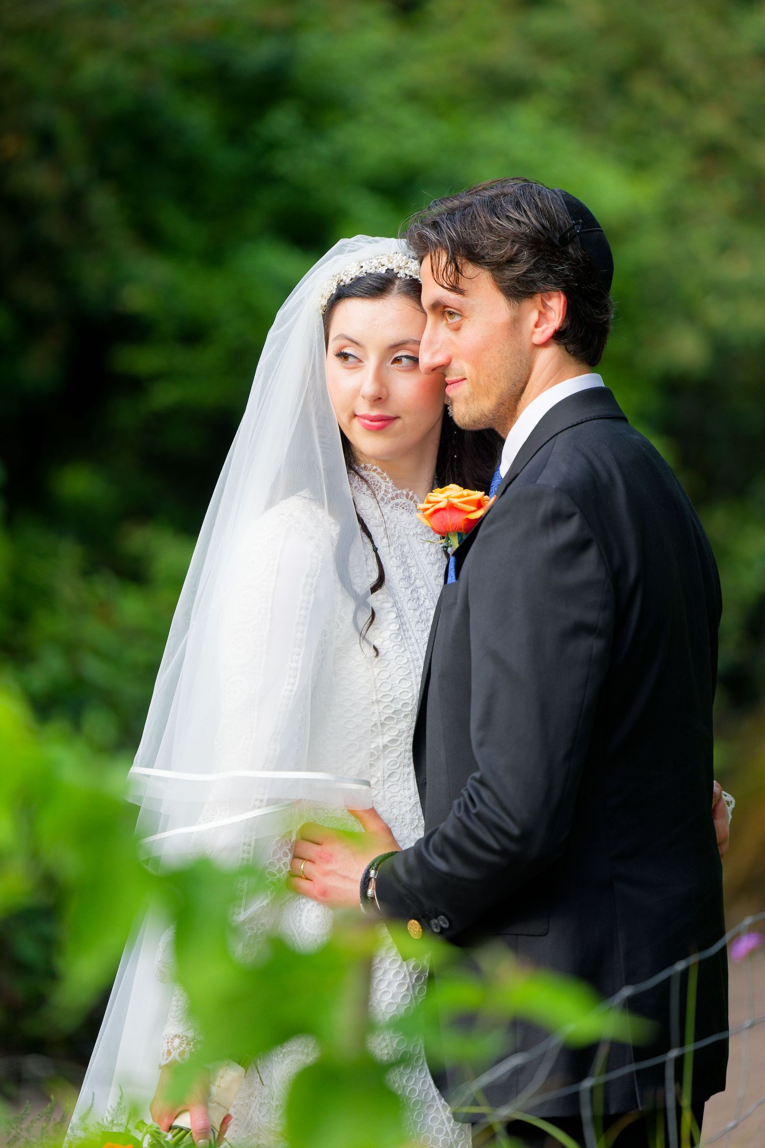 A bride and groom share an intimate moment in their wedding attire against a lush green garden background.