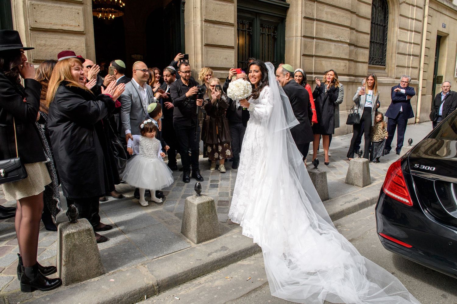 A wedding celebration outside a historic building with a bride in a flowing white gown and veil surrounded by guests.