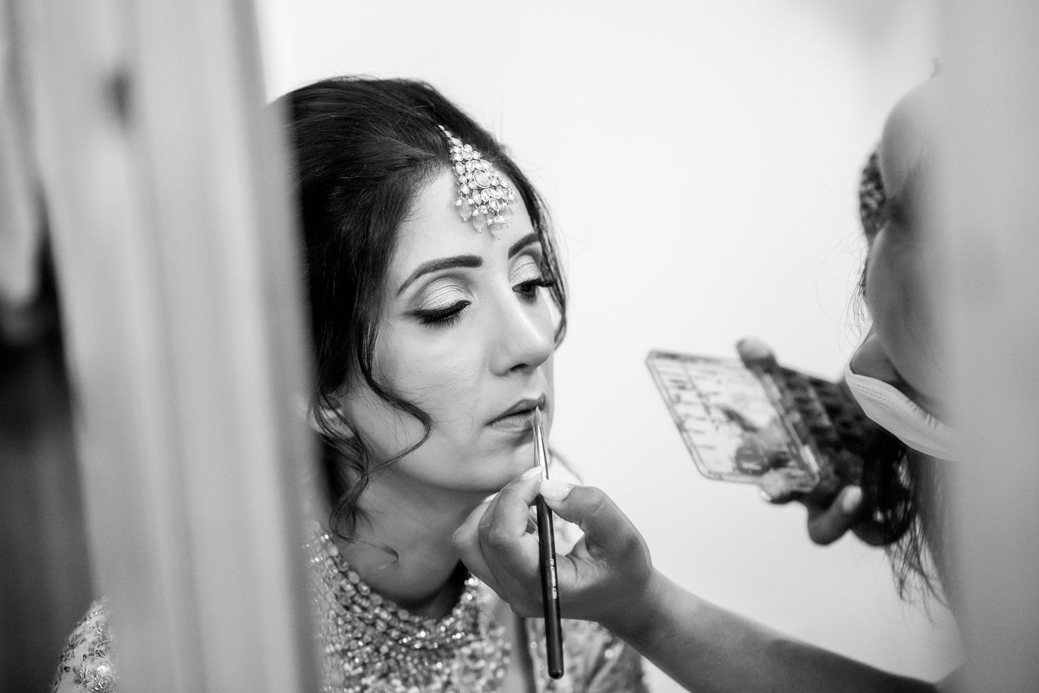 Black and white photo of a bride getting her makeup applied while wearing traditional Indian bridal jewelry and accessories.
