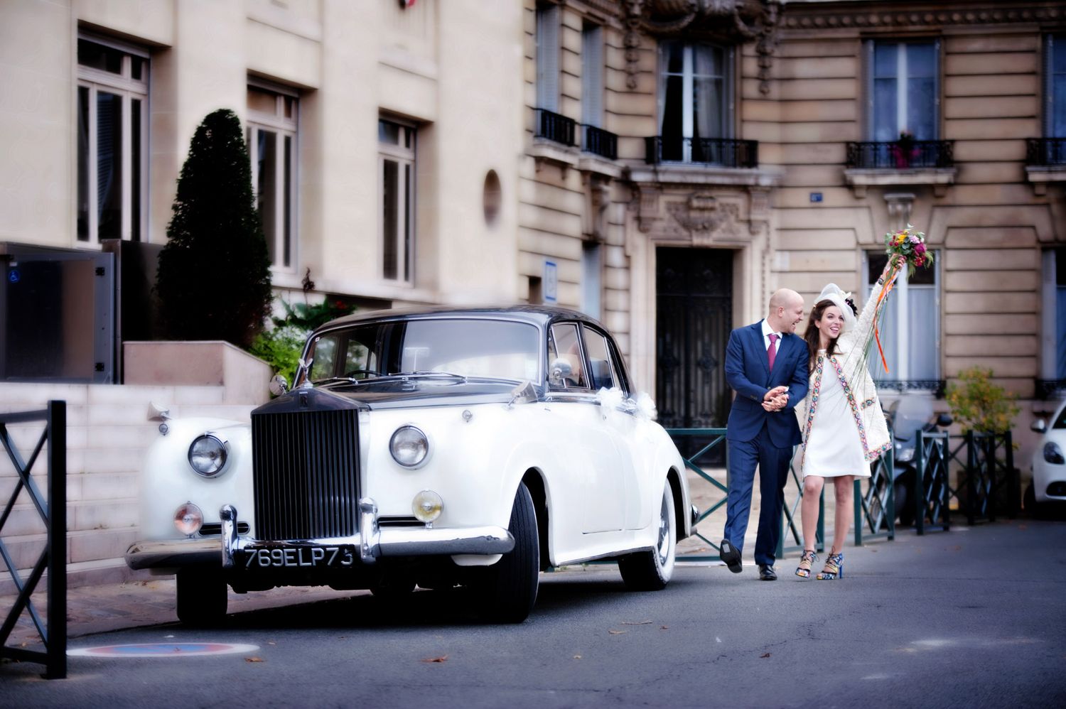 Elegant bride and groom walking beside a classic white Rolls-Royce on a Parisian street.