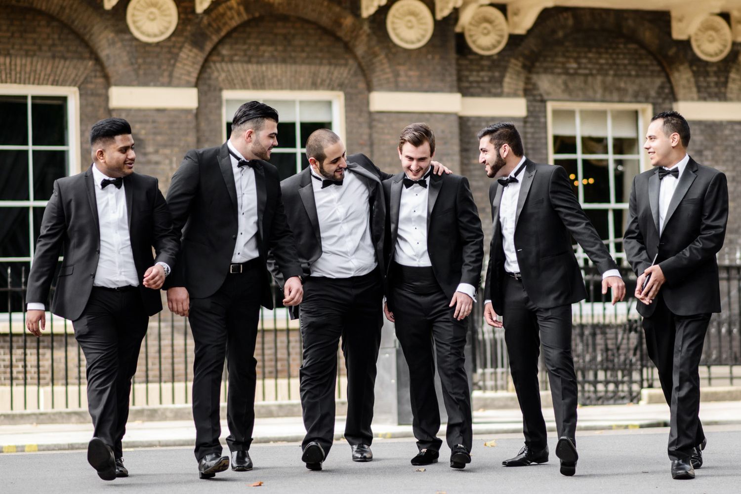 Six men in black tuxedos and bow ties walking and laughing together outside a brick building.