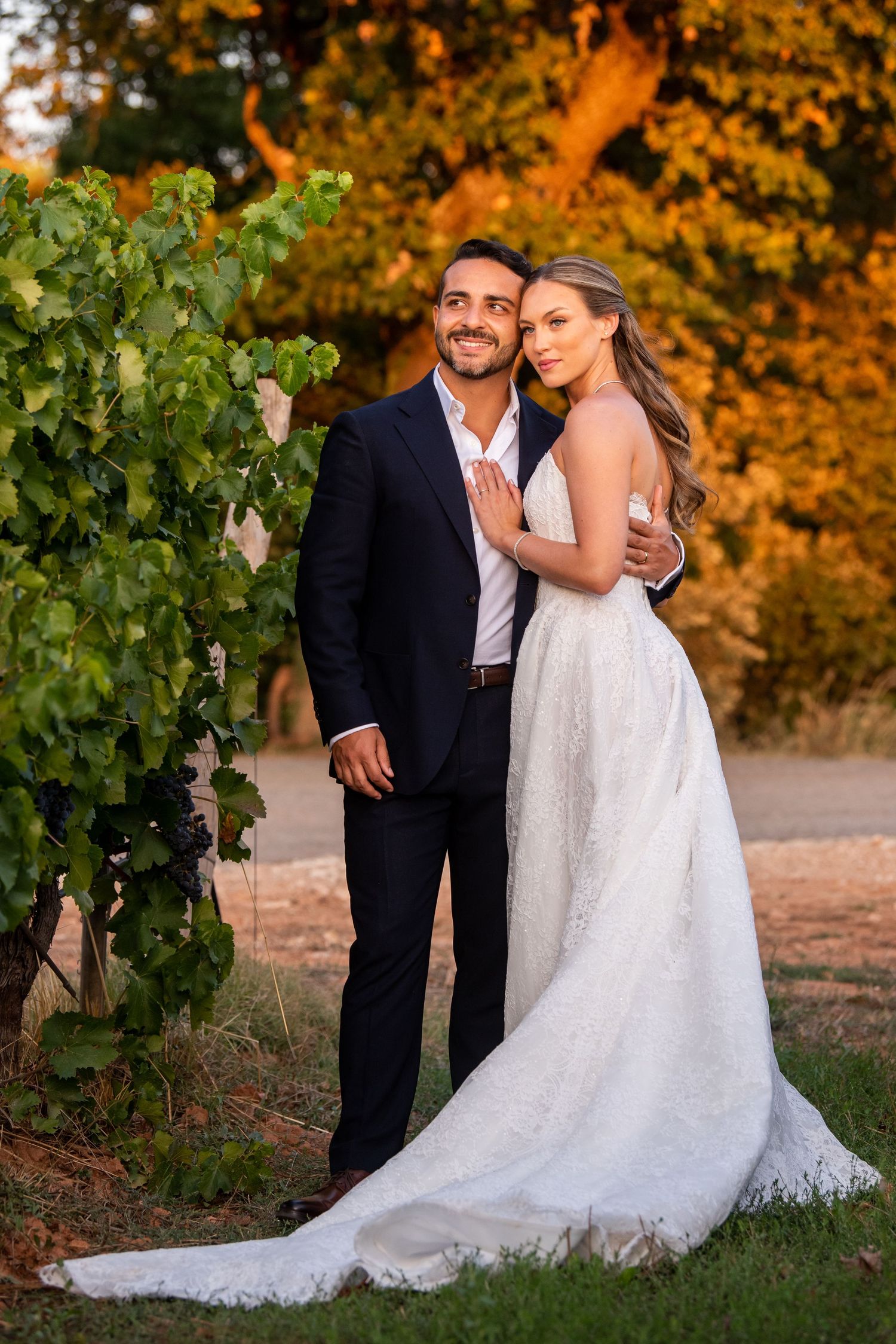Bride and groom posing together in a vineyard at golden hour, bride in white gown, groom in navy suit.