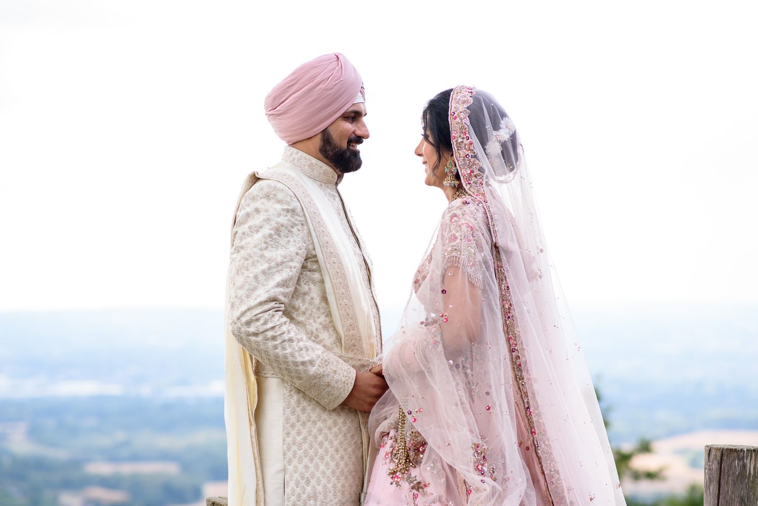 Indian bride and groom in traditional pink and ivory wedding attire holding hands outdoors with scenic backdrop.