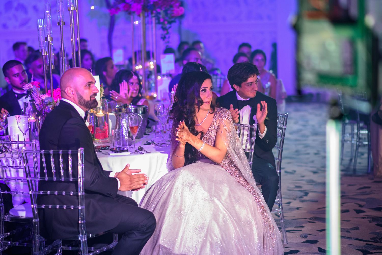 Wedding reception guests dancing and celebrating under purple lighting in an elegant ballroom setting.