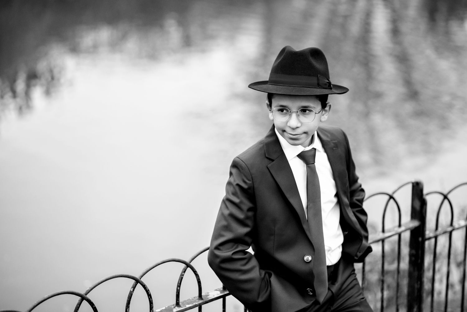 A black and white image shows someone in a suit and fedora hat walking along a wrought iron fence.