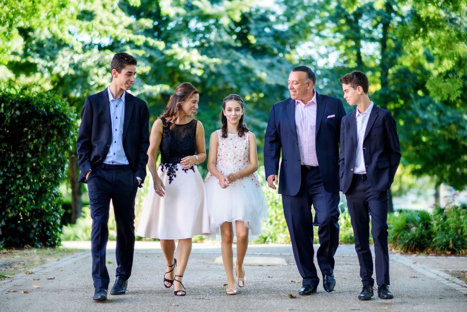 Wedding party walking together on a tree-lined path in formal attire during an outdoor summer celebration.