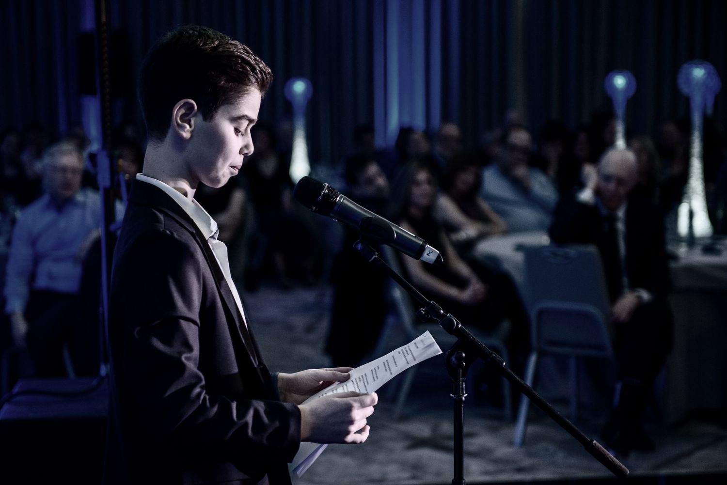 A person reads from a document at a podium in a dimly lit room during a formal event.