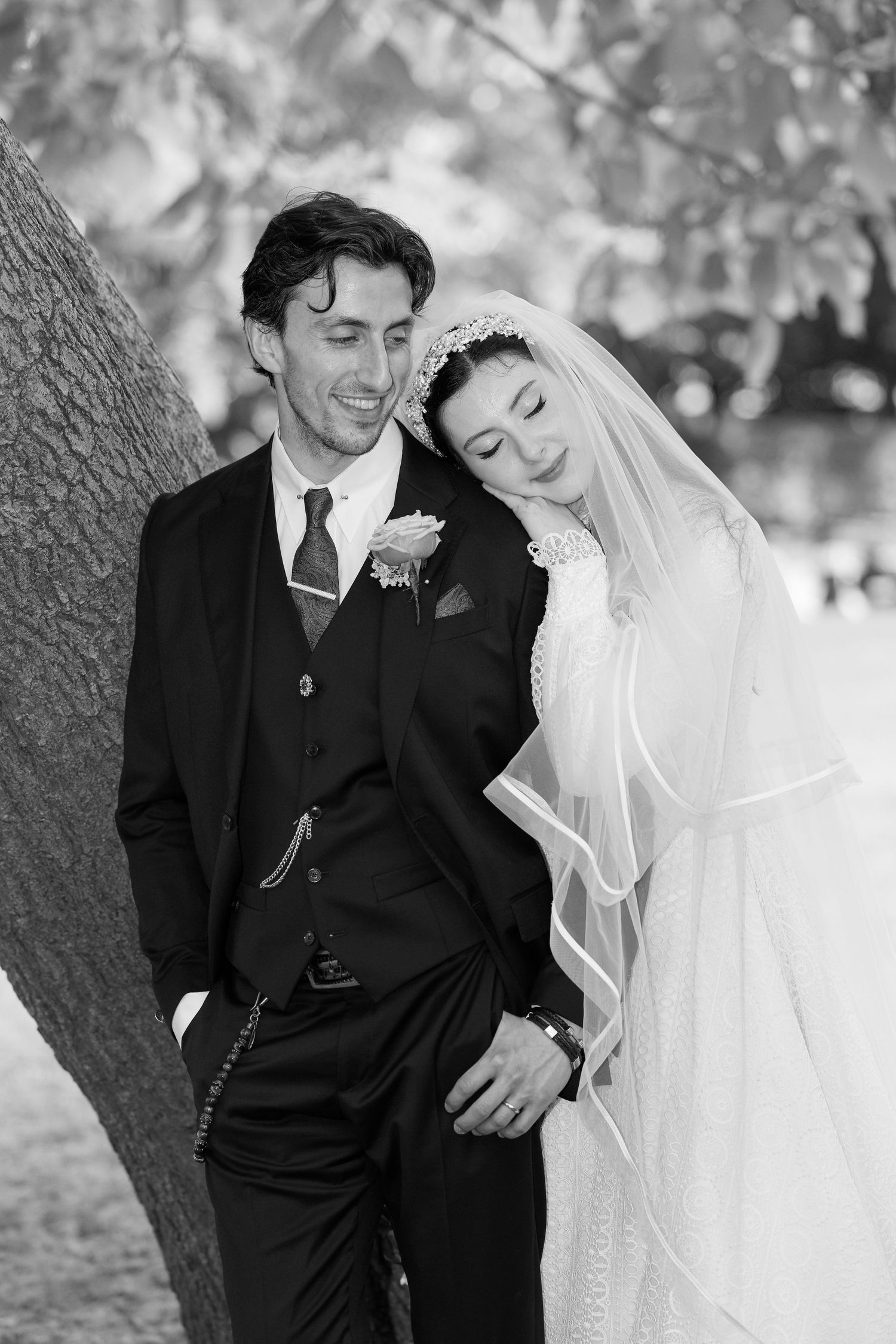 A joyful black and white wedding portrait shows a couple laughing together while leaning against a tree.