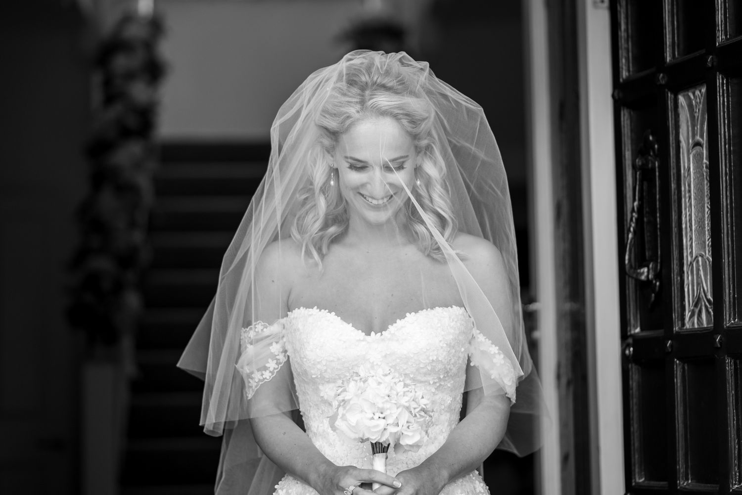 A bride in a white strapless wedding gown stands in a doorway holding a white bouquet.