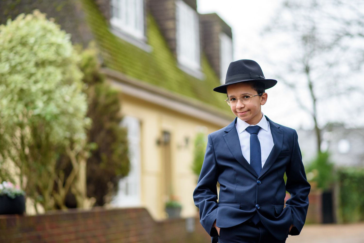 A person in a navy suit and black hat poses confidently in front of a yellow building.