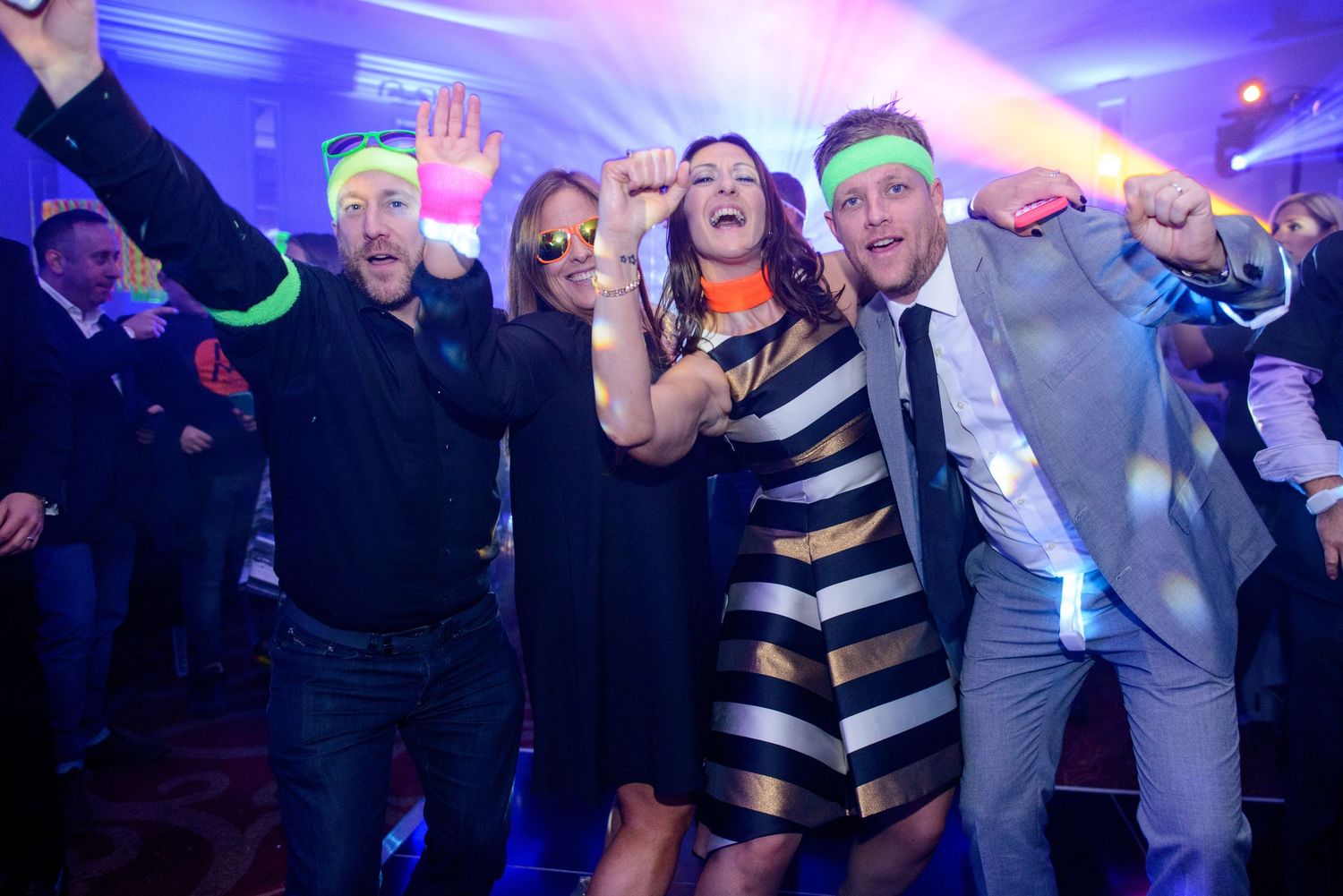 Party guests wearing neon glow sticks dance and celebrate on a dance floor with blue lighting effects.