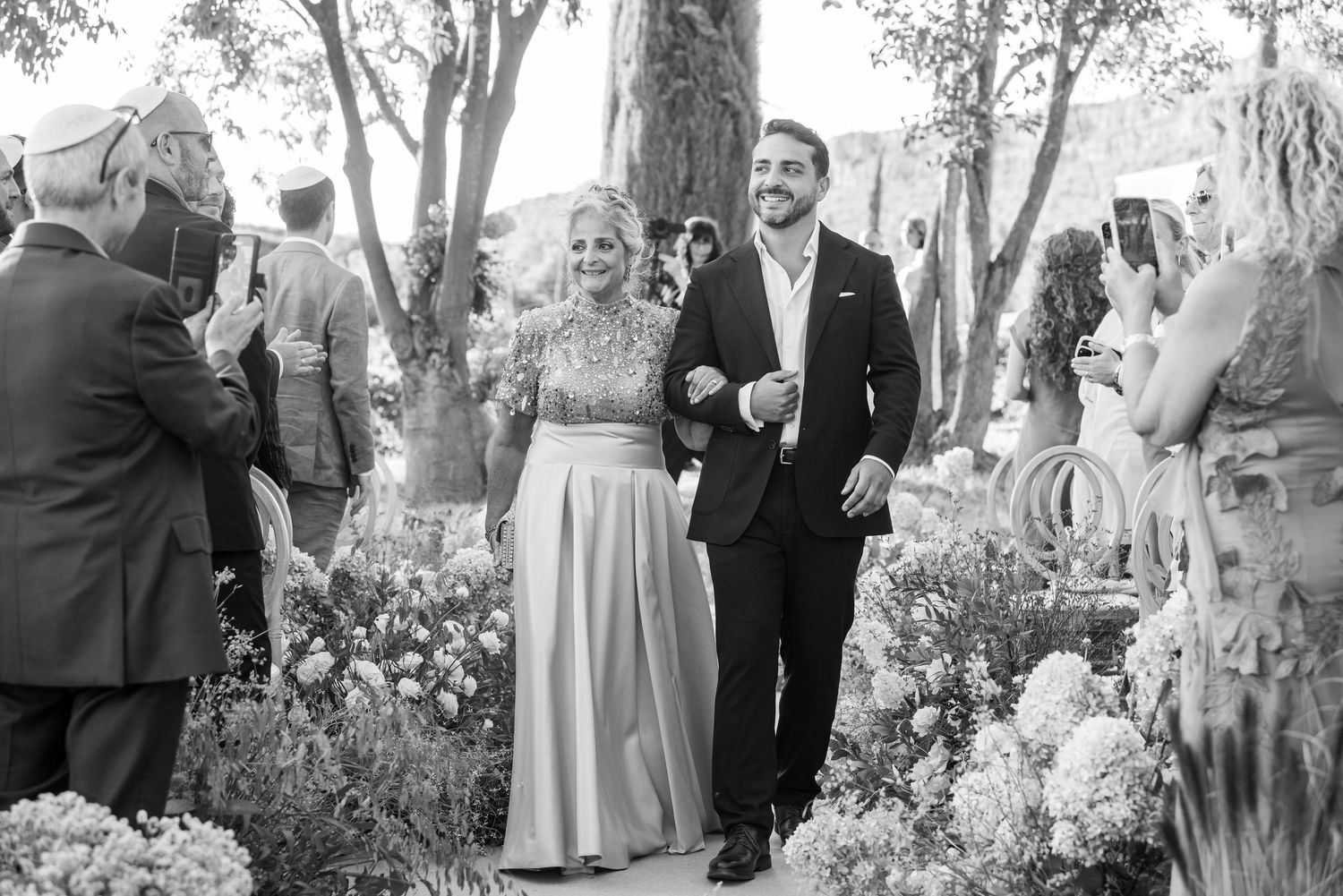 A black and white wedding photo showing a couple walking through a garden with guests and floral decorations.