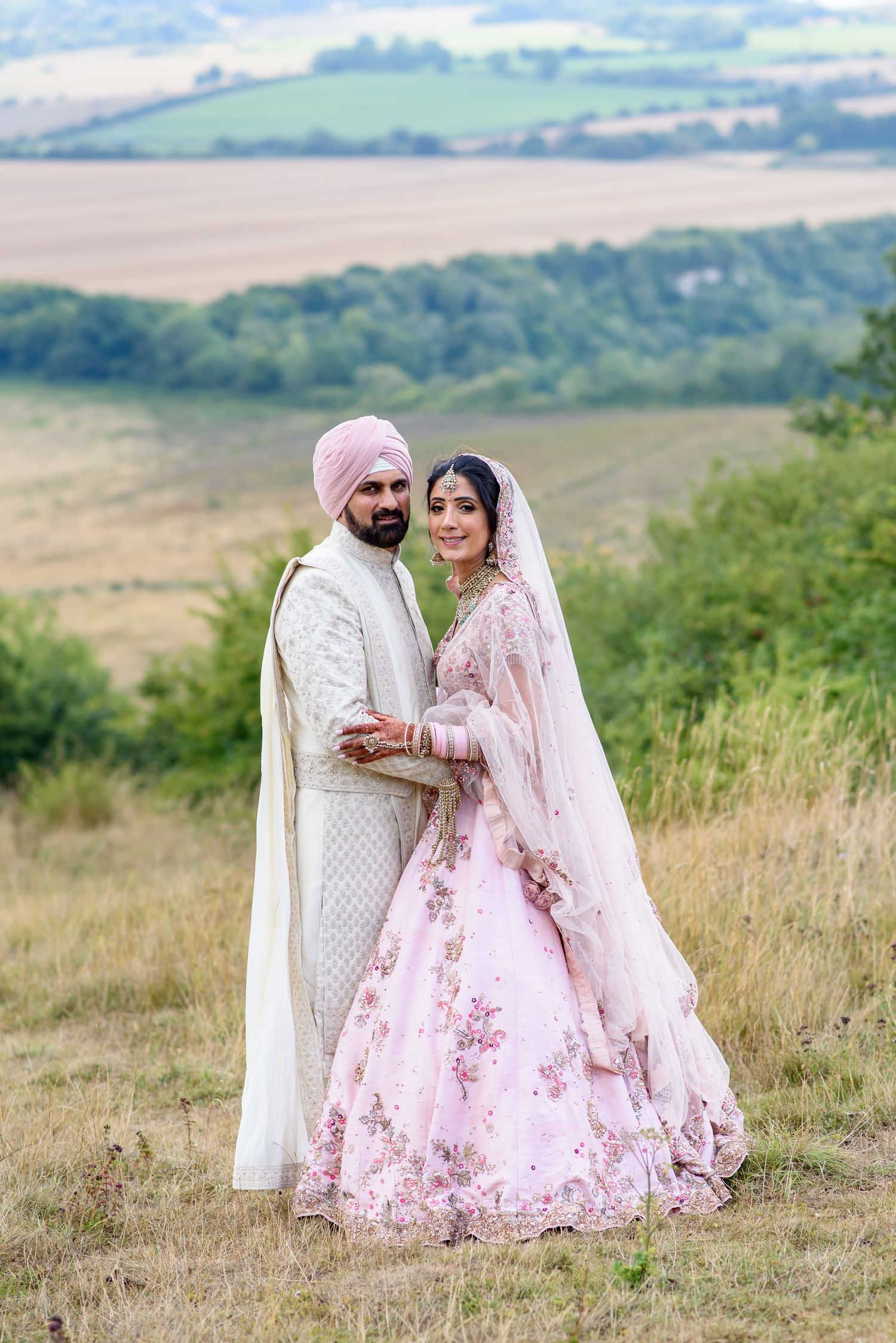 Sikh couple in traditional wedding attire, groom in white sherwani and pink turban, bride in floral pink lehenga outdoors.