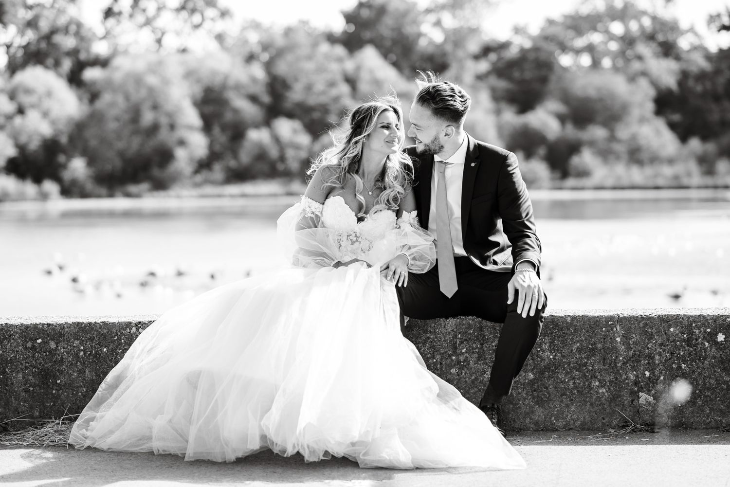 Black and white photo of a bride and groom sitting together on a stone wall, sharing an intimate moment outdoors.