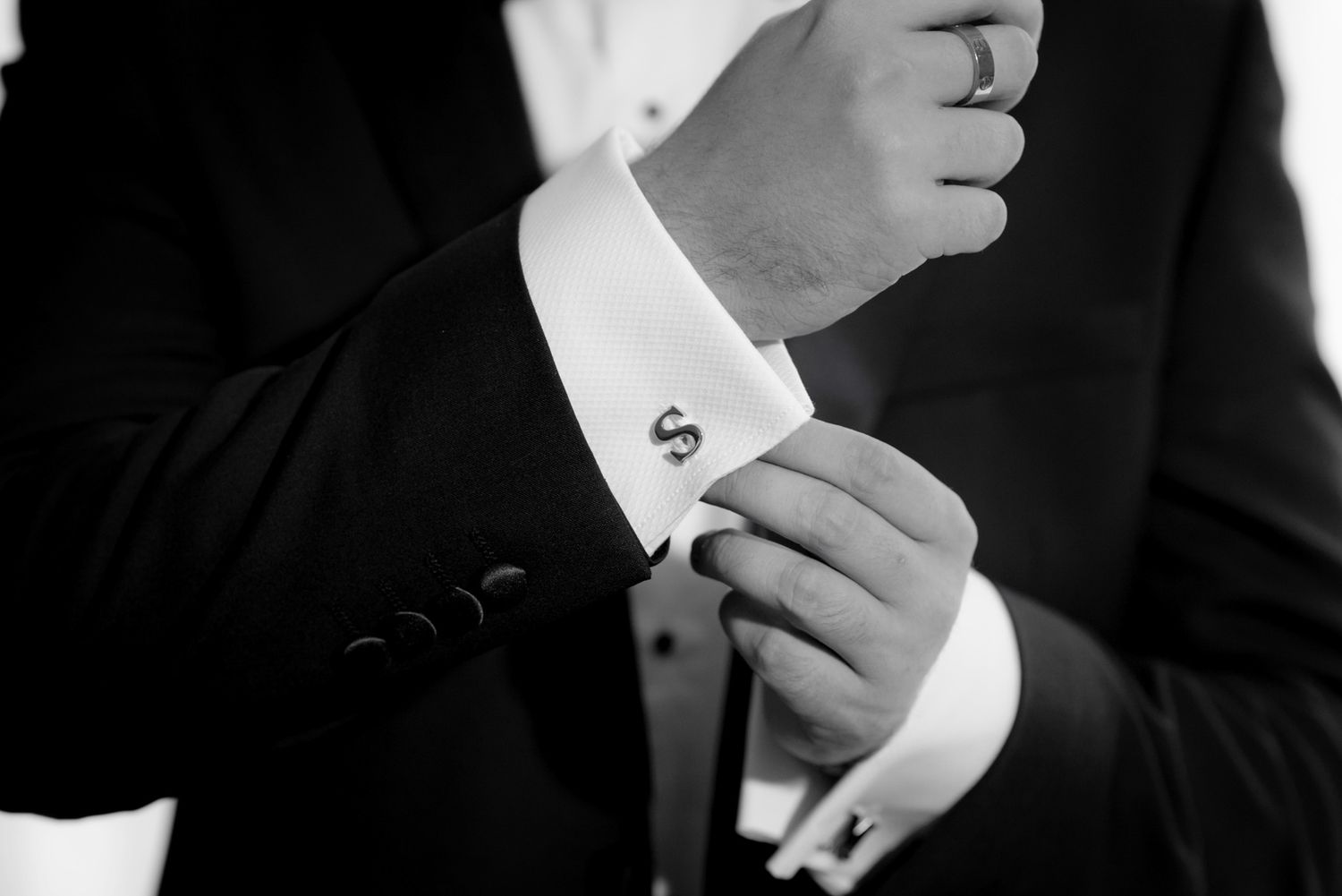 Black and white photo of a groom fastening monogram S cufflinks on his suit sleeve before the wedding ceremony.