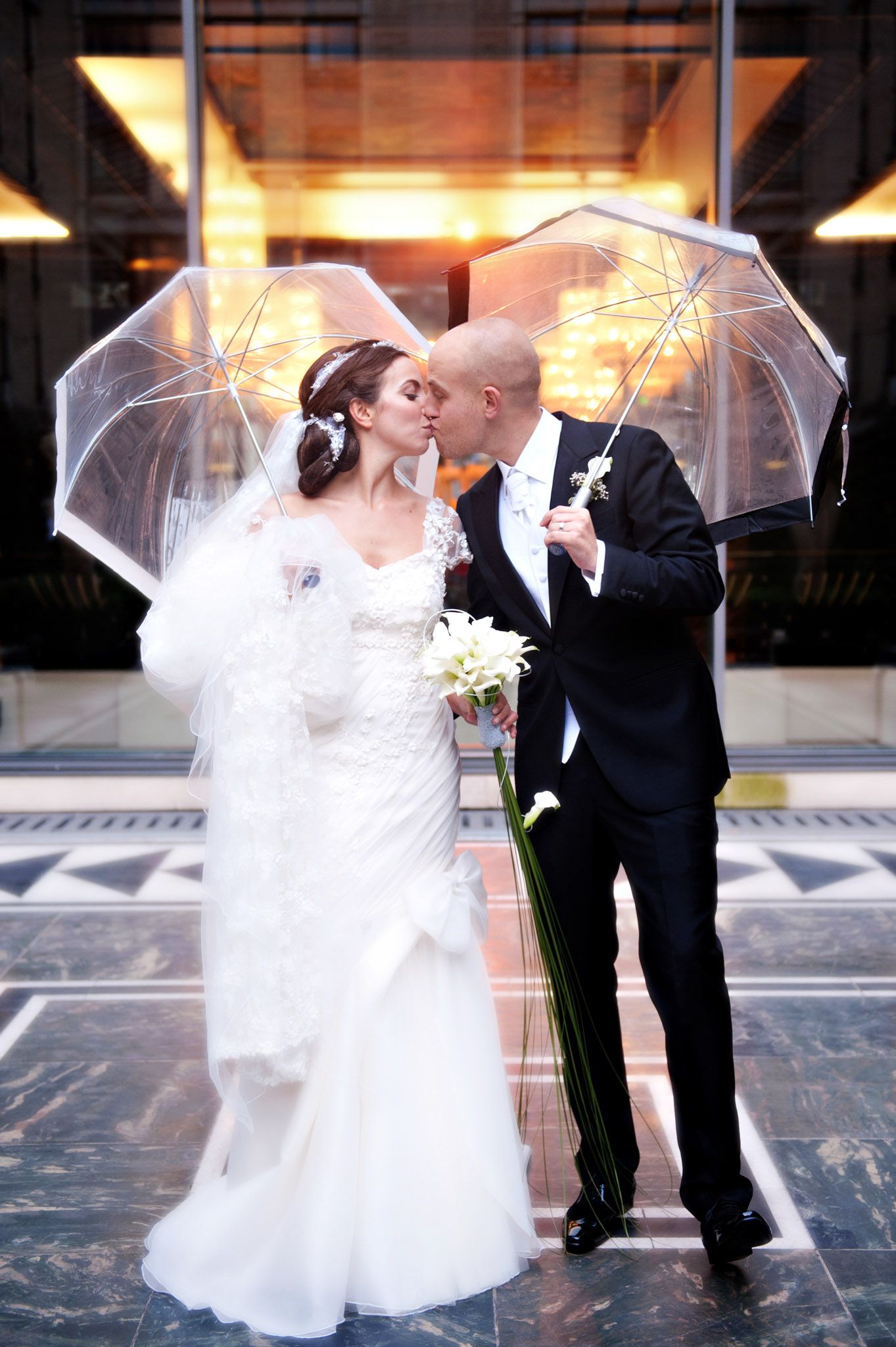 Bride and groom kissing under clear umbrellas on their wedding day, bride in white gown with veil holding calla lilies.