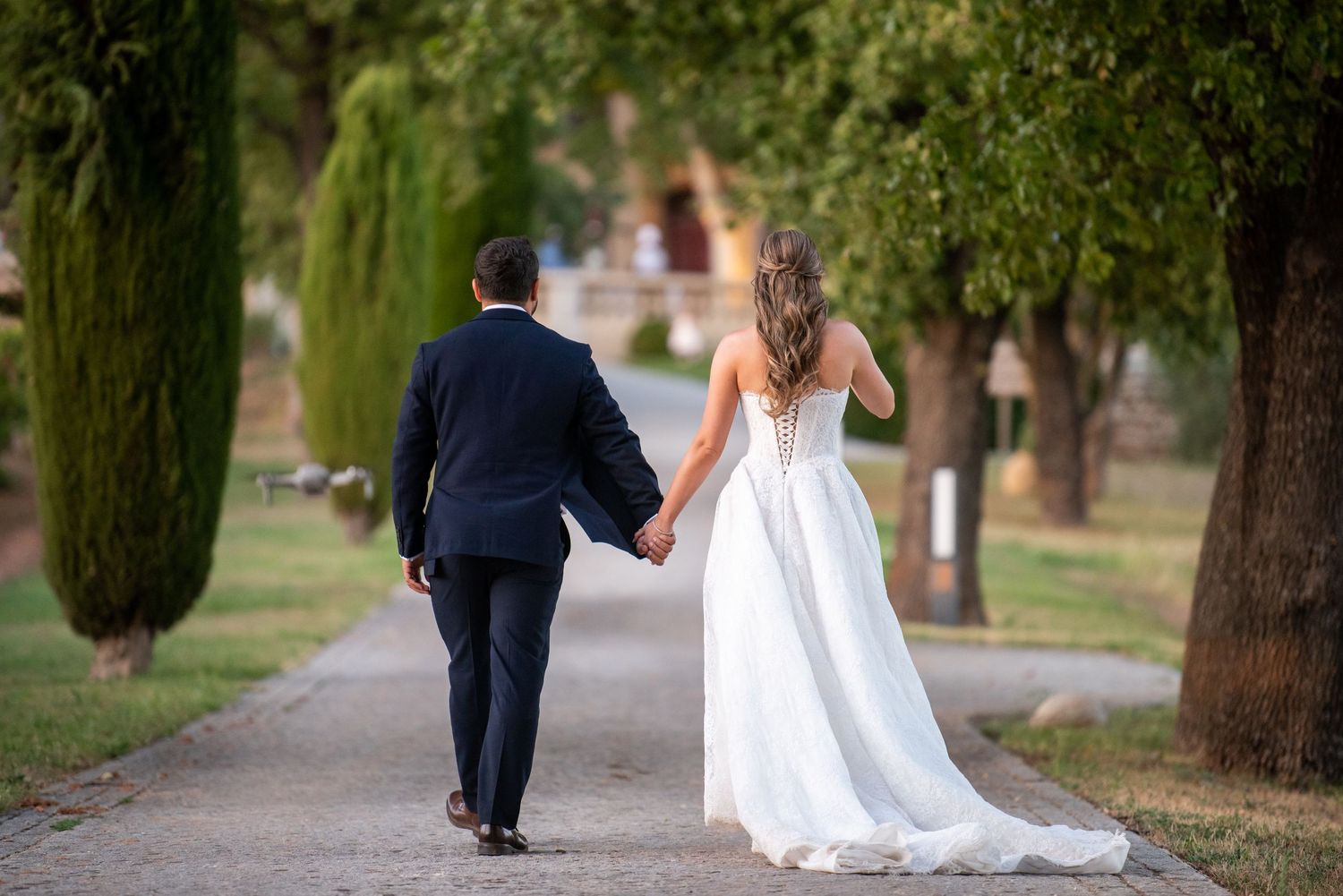 Bride and groom walking hand in hand down a tree-lined path, bride in white gown, groom in navy suit.