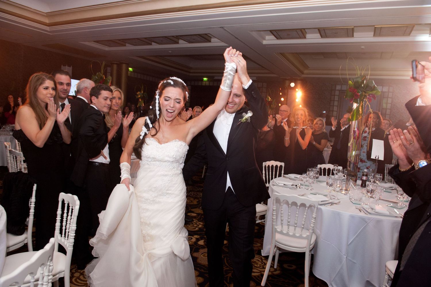 Wedding guests dance and celebrate during an evening reception in a dimly lit ballroom with white table settings.