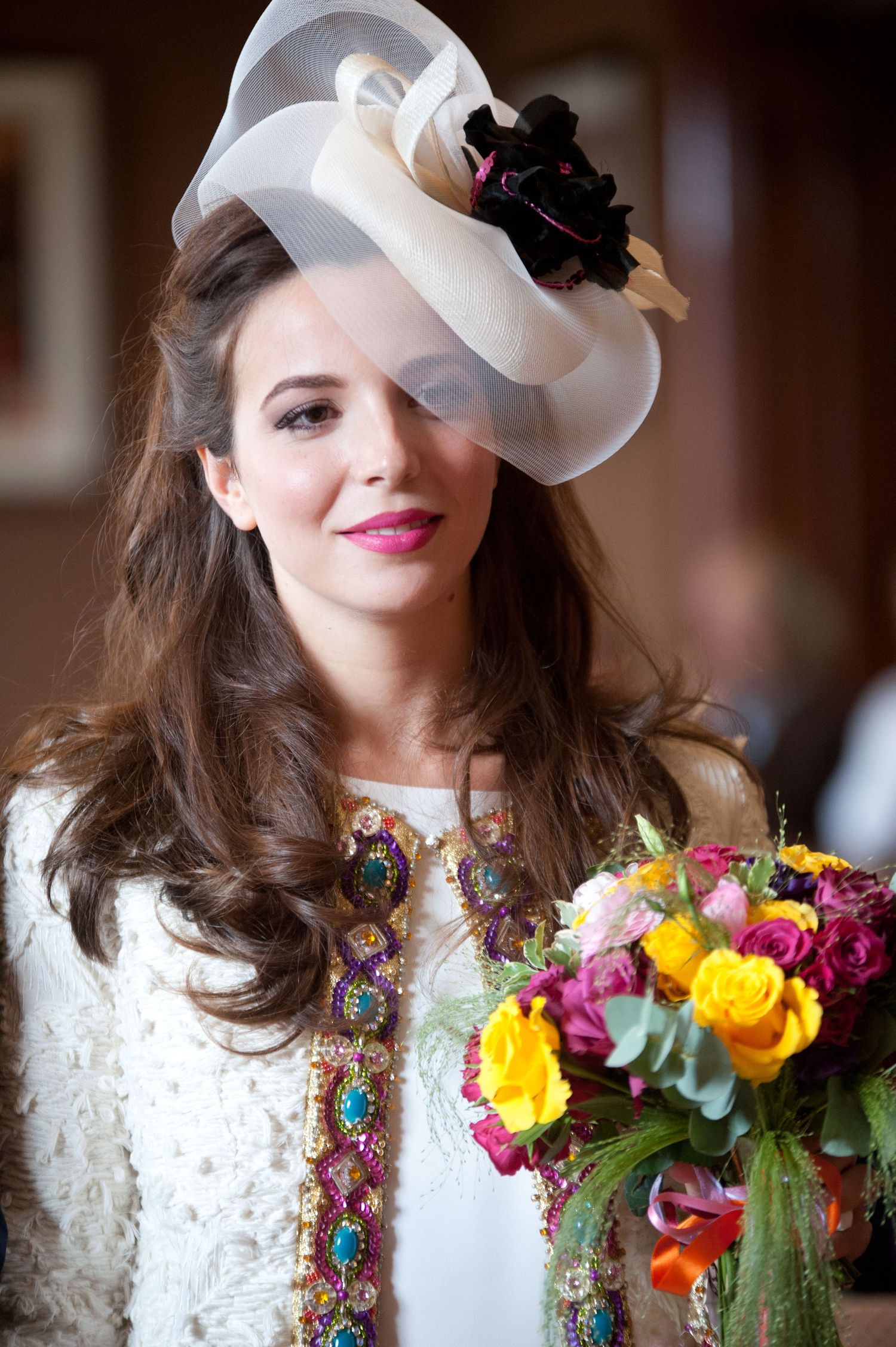A person in white lace wearing a vintage-style hat and colorful floral necklace smiles at the camera.