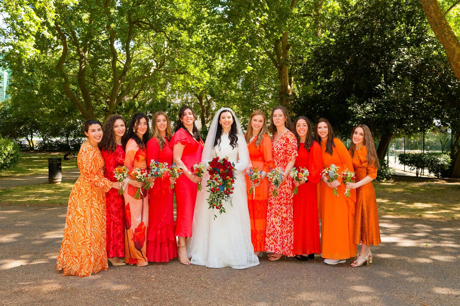 Wedding party in vibrant orange and red floral dresses poses outdoors in a sunny garden setting with the bride in white.