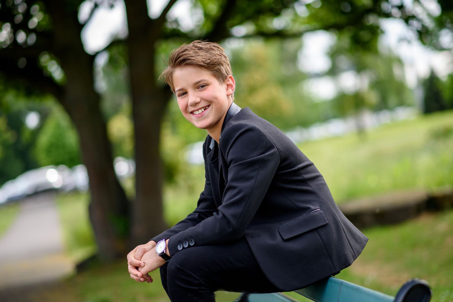 A person in a black suit jacket poses outdoors on a sunny day with trees and greenery in the background.