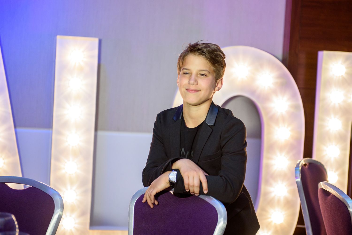 A performer sits on a couch in front of illuminated marquee lights on a purple-lit stage set.