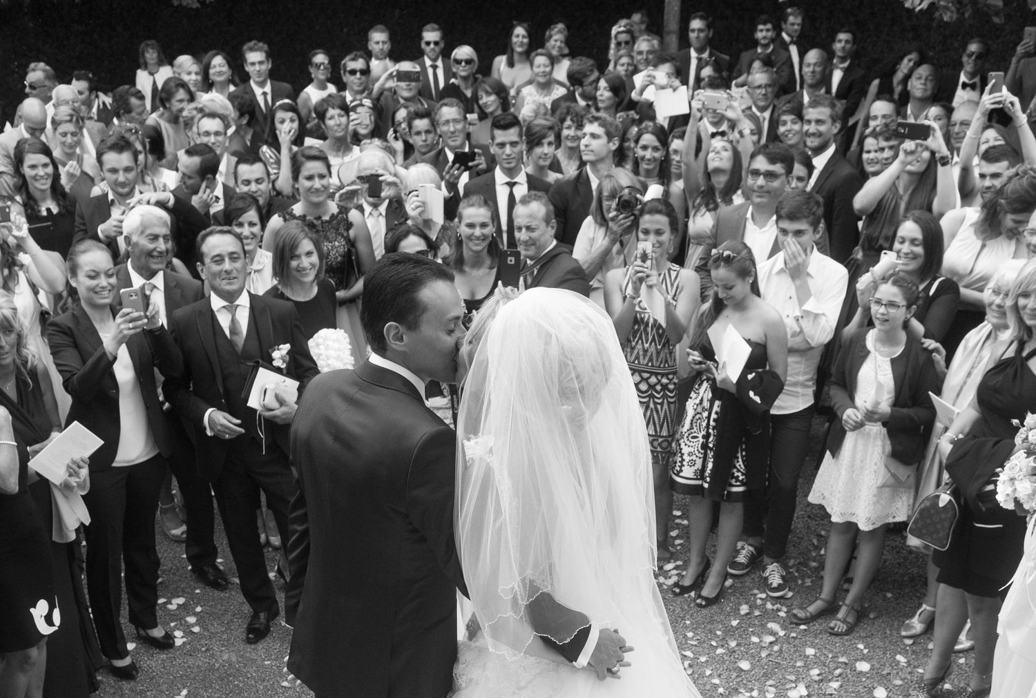 Bride and groom sharing a kiss surrounded by wedding guests in a classic black and white photo.
