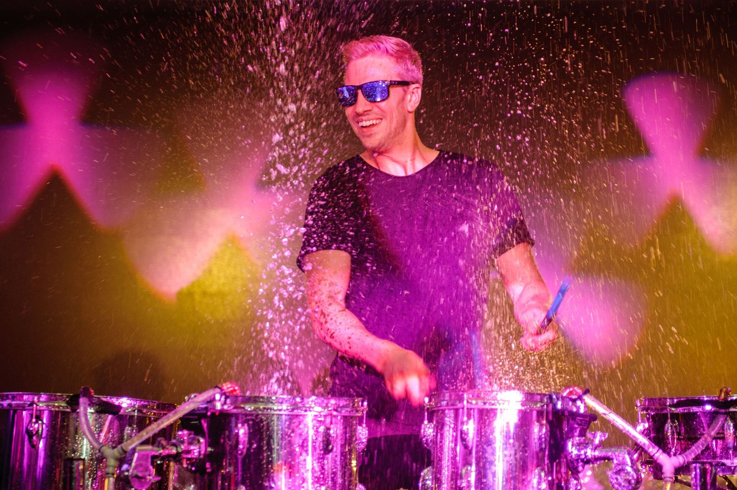 Drummer performs on stage surrounded by pink and purple lighting effects while water droplets spray through the air.