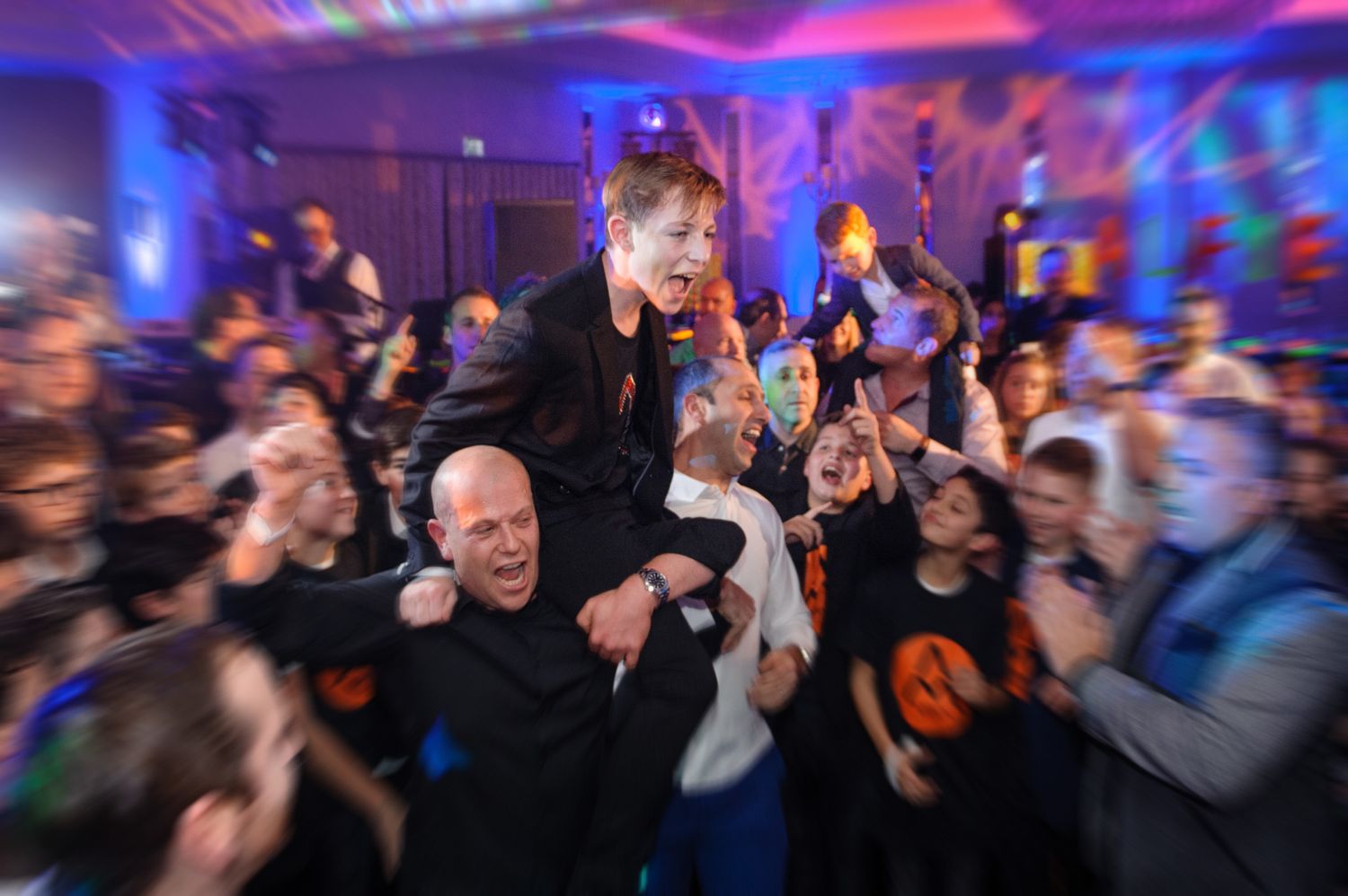Energetic party crowd dancing and celebrating under vibrant blue and purple lighting at a nightclub event.