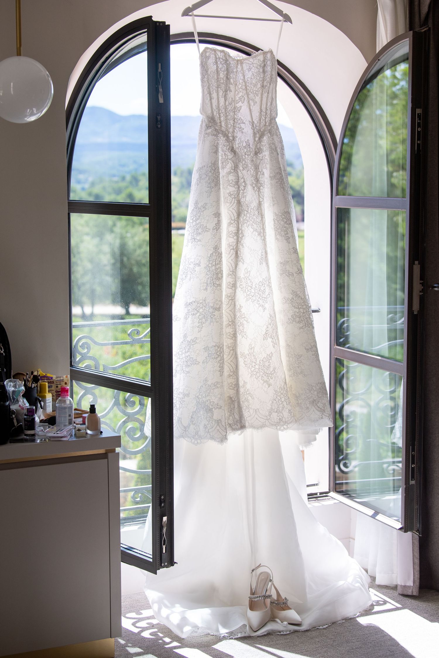 A white wedding dress hangs elegantly by a window, showcasing its delicate lace details and flowing silhouette in natural light.