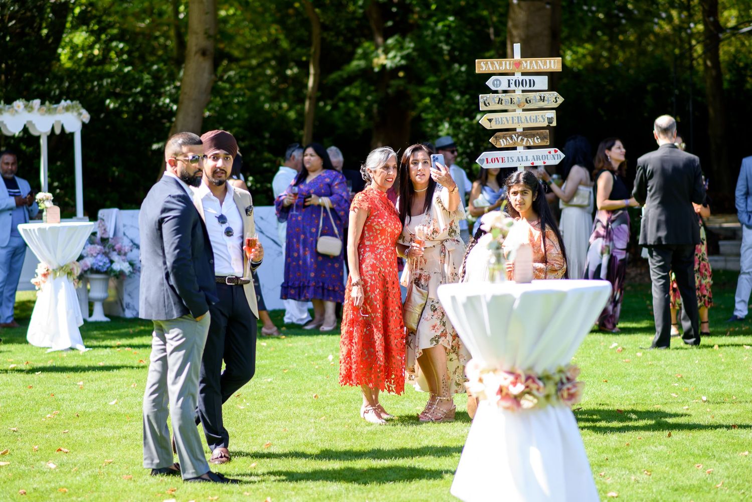 Wedding guests gather outdoors for a reception on a sunny day with white cocktail tables and green lawn.