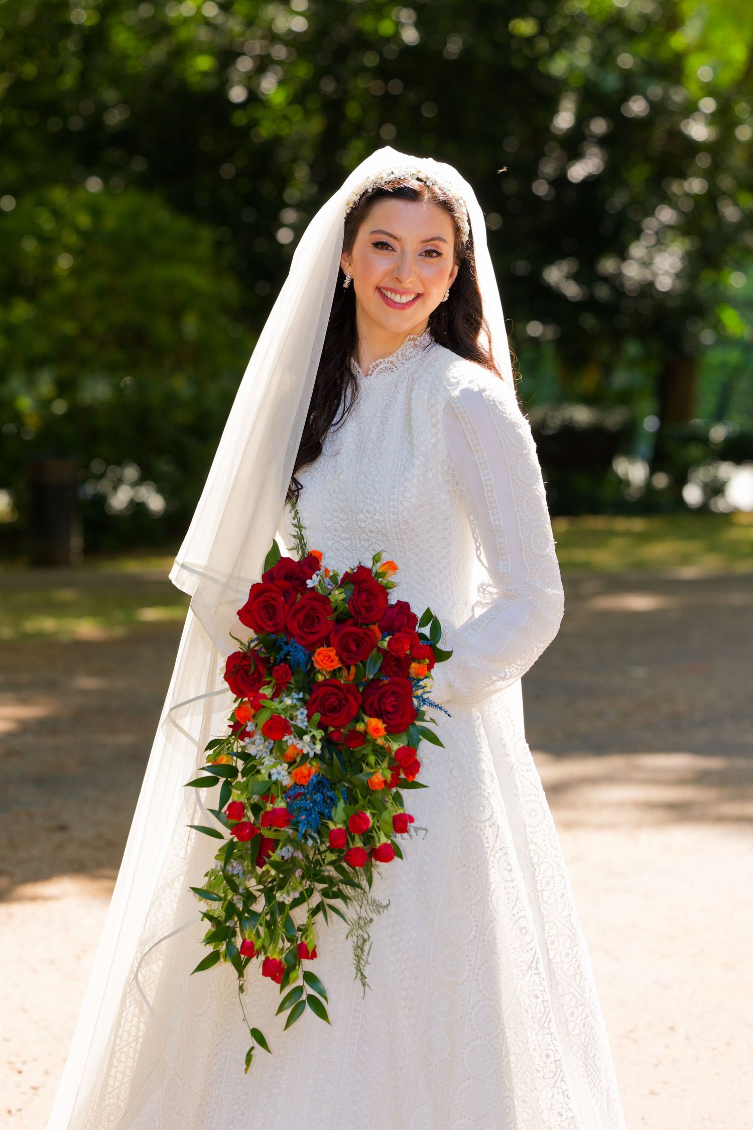 A bride in a white lace dress holds a vibrant red and blue bouquet while posing outdoors in a garden setting.
